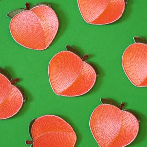 Könnte beinhalten: Ein Set mit zehn Pfirsich-farbenen Filz-Pfirsich-Patches. Die Patches sind mit einem detaillierten Design bestickt und haben eine leicht erhabene Textur.