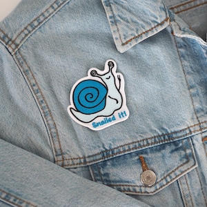 Puede incluir: Una chaqueta vaquera azul con un parche bordado de un caracol blanco y azul que dice "Snailed it!"