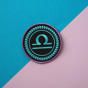 Op de afbeelding: Een geborduurde zwarte en turquoise patch met het astrologische symbool voor Weegschaal, omringd door een paarse en turquoise krans.