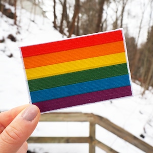 Può includere: Un cerotto di bandiera arcobaleno pride con sei strisce orizzontali nei colori rosso, arancione, giallo, verde, blu e viola. Il cerotto è fatto di tessuto e ha un bordo bianco.
