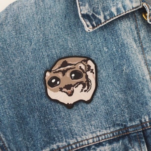 Pode incluir: Um remendo bordado marrom e branco de um pequeno animal com olhos grandes, nariz preto e boca rosa. O remendo é costurado em uma jaqueta jeans azul.