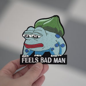 May include: An embroidered iron-on patch featuring a blue and green cartoon frog with the text "FEELS BAD MAN" on a black background.