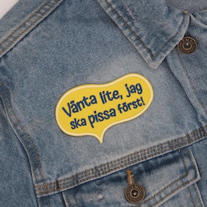 Op de afbeelding: Een gele spraakballon-patch genaaid op een blauw denim jack. De patch zegt "Vänta lite, jag ska pissa först!" in blauwe tekst.