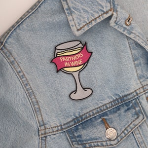 Può includere: Una giacca di jeans blu chiaro con una toppa ricamata a forma di bicchiere di vino bianco con la scritta "Partners in Wine" in giallo su uno stendardo rosso.