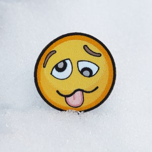 Può includere: Emoticon ricamato giallo con la lingua che sporge e gli occhi chiusi. L'emoticon ha un sopracciglio marrone su ogni lato.
