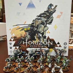 Könnte beinhalten: Ein Brettspielkarton mit dem Titel "Horizon Zero Dawn: The Board Game" mit einer Illustration einer Roboter-Kreatur. Das Spiel enthält Miniaturfiguren von Charakteren und Kreaturen, die in verschiedenen Farben bemalt und auf kreisförmigen Sockeln montiert sind.