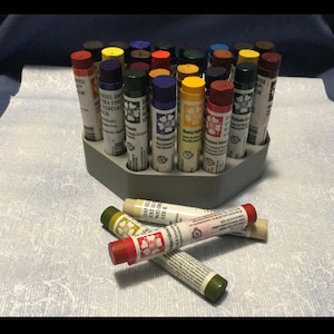 May include: A hexagonal gray holder with 20 tubes of watercolor paint. The tubes are arranged in a circle around the holder. The tubes are labeled with the brand name "Daniel Smith" and the color name. The tubes are a variety of colors, including red, blue, green, yellow, and brown.