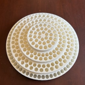 May include: A white, circular object with a series of concentric rings. Each ring has a pattern of evenly spaced holes. The object is made of a light-colored material and sits on a dark brown surface.