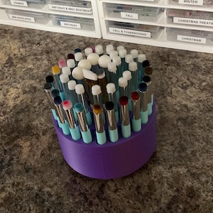 May include: A purple makeup brush holder filled with various makeup brushes. The brushes have silver handles, light blue bases, and different colored brush heads. The holder is on a brown surface, with labeled storage containers in the background.