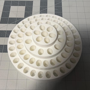 May include: A white, multi-tiered, circular organizer with numerous oval-shaped holes. The organizer is designed to hold small items, likely for crafting or hobbies. The design features concentric rings, creating a visually appealing and functional storage solution.