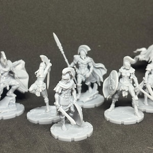 May include: A collection of gray miniature figurines depicting ancient warriors. The figures include soldiers with shields, swords, spears, and a flag. The miniatures are on circular bases and are set against a dark background.