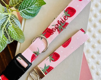 May include: A pink dog collar with a strawberry print and the name "Sophie" embroidered on it. The collar is paired with a matching pink leash with a silver key ring.