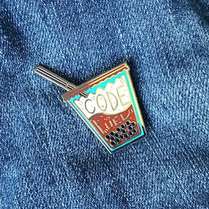 Peut inclure: Épinglette en émail colorée en forme de tasse de thé aux perles, avec les mots "CODE FUEL" en blanc et marron. L'épinglette a une paille argentée et est posée sur un fond en jean bleu.