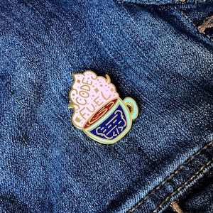 Peut inclure: Épinglette en émail doré représentant une tasse de café avec un tourbillon rose et blanc sur le dessus. La tasse a une bordure bleue et les mots "CODE FUEL" sont écrits sur le tourbillon.