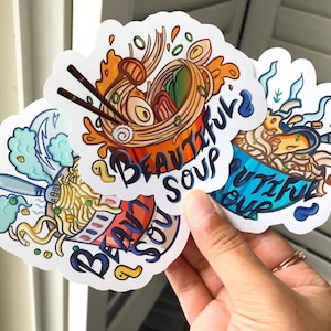 May include: A hand holds a stack of die-cut stickers featuring colorful illustrations of ramen bowls. Each sticker has the words "Beautiful Soup" in a whimsical font. The ramen bowls are depicted with various ingredients and styles.