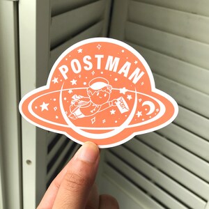 K&ouml;nnte beinhalten: Ein pfirsichfarbener Aufkleber mit einem wei&szlig;en Umriss eines Comic-Charakters, der mit einem Brief in der Hand durch den Weltraum fliegt. Der Aufkleber sagt "POSTMAN" in einem wei&szlig;en Kreis mit wei&szlig;en Sternen.