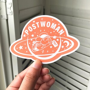 K&ouml;nnte beinhalten: Ein pfirsichfarbener Aufkleber mit einem wei&szlig;en Umriss eines Planeten und einer Cartoonfigur, die im Weltraum fliegt. Die Figur h&auml;lt einen wei&szlig;en Umschlag mit dem Wort "POST" darauf. Der Text "POSTWOMAN" ist um den Planeten herum geschrieben.