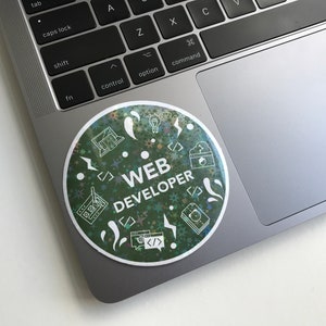 Pode incluir: Um autocolante redondo num port&aacute;til com as palavras "WEB DEVELOPER" em branco. O autocolante tem um fundo verde com &iacute;cones brancos de c&oacute;digo inform&aacute;tico, um ecr&atilde; de computador e uma l&acirc;mpada. O autocolante est&aacute; num port&aacute;til prateado.
