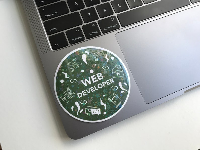 Pode incluir: Um autocolante redondo num port&aacute;til com as palavras "WEB DEVELOPER" em branco. O autocolante tem um fundo verde com &iacute;cones brancos de c&oacute;digo inform&aacute;tico, um ecr&atilde; de computador e uma l&acirc;mpada. O autocolante est&aacute; num port&aacute;til prateado.