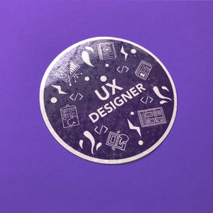Op de afbeelding: Een ronde sticker met een paarse achtergrond en een wit ontwerp. De sticker zegt "UX Designer" en bevat pictogrammen die gerelateerd zijn aan user experience design, zoals een computerscherm, een notitieblok en een gloeilamp.