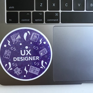 Op de afbeelding: Een ronde, paarse sticker met witte pictogrammen en de tekst "UX DESIGNER". De pictogrammen omvatten een klembord, een notitieboek, een grafiek, een telefoon en een bliksem.