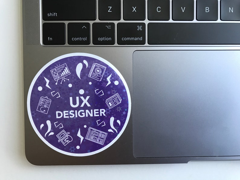 Op de afbeelding: Een ronde, paarse sticker met witte pictogrammen en de tekst "UX DESIGNER". De pictogrammen omvatten een klembord, een notitieboek, een grafiek, een telefoon en een bliksem.