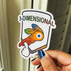 May include: A sticker with the text "3-DIMENSIONAL" in rainbow colors. The sticker features a stylized orange, yellow, and blue design with a white hand pointing at it. The sticker is held by a person.