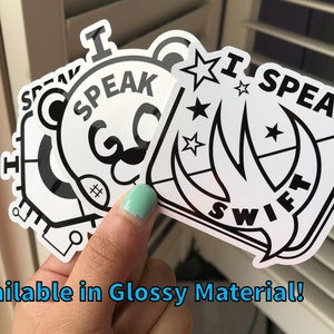 May include: A hand holds several white stickers with black text and designs. The stickers say "I SPEAK SWIFT" and "I SPEAK" with a bear face. The text "Available in Glossy Material!" is at the bottom.