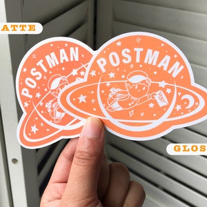 K&ouml;nnte beinhalten: Zwei orangefarbene Aufkleber mit wei&szlig;em Rand. Die Aufkleber zeigen eine Comicfigur in einem Raumanzug, die einen Brief mit der Aufschrift "Post" h&auml;lt. Der Text "POSTMAN" ist in einem wei&szlig;en Kreis um die Figur geschrieben. Ein Aufkleber ist mit "Matt" und der andere mit "Glanz" beschriftet.