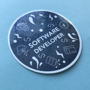 Może przedstawiać: Okrągła naklejka z granatowym tłem i białymi ilustracjami symboli i narzędzi kodowania. Tekst "SOFTWARE DEVELOPER" jest biały, z białą obw&oacute;dką. Naklejka znajduje się na jasnoniebieskiej powierzchni.