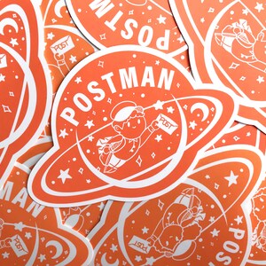 K&ouml;nnte beinhalten: Ein Stapel orangefarbener und wei&szlig;er Aufkleber mit einem Comic-Astronauten, der Post liefert. Die Aufkleber zeigen den Text "POSTMAN" und einen Halbmond.