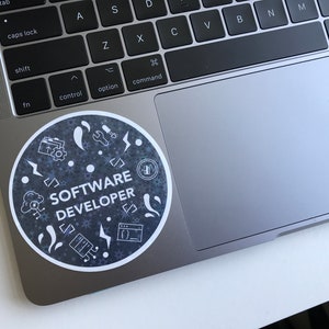 Może przedstawiać: Okrągła naklejka z białym napisem "SOFTWARE DEVELOPER", otoczona białymi ikonami na ciemnoszarym tle. Naklejka znajduje się na srebrnym laptopie.