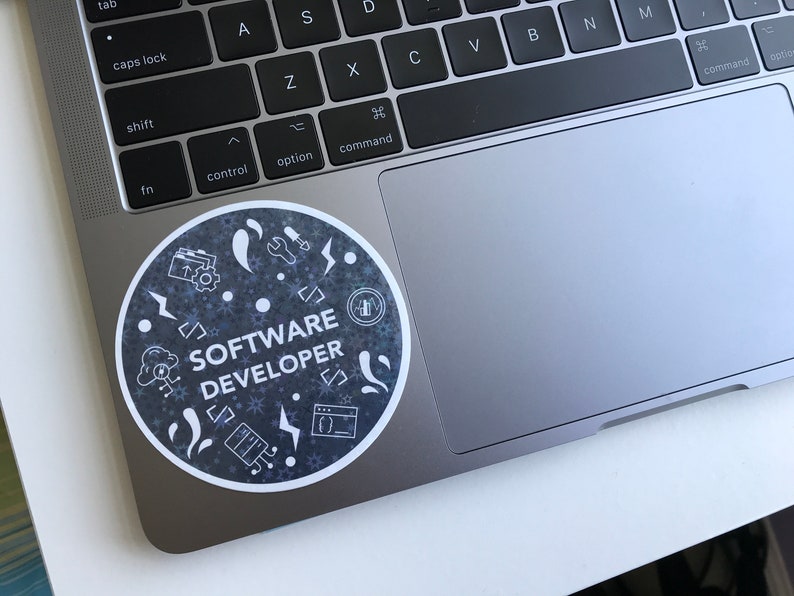 Może przedstawiać: Okrągła naklejka z białym napisem "SOFTWARE DEVELOPER", otoczona białymi ikonami na ciemnoszarym tle. Naklejka znajduje się na srebrnym laptopie.