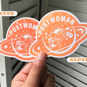 K&ouml;nnte beinhalten: Zwei orangefarbene Aufkleber mit wei&szlig;em Rand. Die Aufkleber zeigen eine Comicfigur, die mit einem Paket, auf dem "POST" steht, durch den Weltraum fliegt. Der Text auf den Aufklebern lautet "POSTWOMAN". Ein Aufkleber ist mit "MATTE" und der andere mit "GLOSSY" beschriftet.