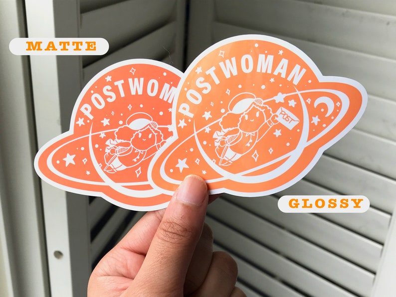 K&ouml;nnte beinhalten: Zwei orangefarbene Aufkleber mit wei&szlig;em Rand. Die Aufkleber zeigen eine Comicfigur, die mit einem Paket, auf dem "POST" steht, durch den Weltraum fliegt. Der Text auf den Aufklebern lautet "POSTWOMAN". Ein Aufkleber ist mit "MATTE" und der andere mit "GLOSSY" beschriftet.