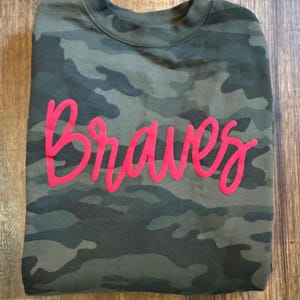May include: A camouflage-patterned sweatshirt with the word "Braves" in bright pink script across the front. The sweatshirt is folded and rests on a wooden surface. The camouflage design features shades of green, brown, and black.
