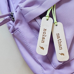 May include: A light purple backpack with a zippered pocket, featuring two wooden tags. The tags are engraved with the names "nolan" and "nathan", each accompanied by a lightning bolt symbol. The tags are attached with neon green cords.