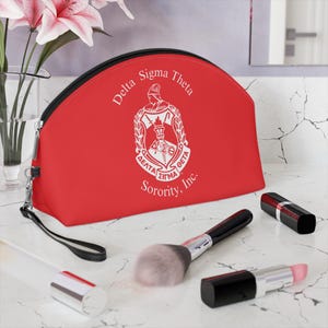 May include: A red cosmetic bag with a black zipper and wrist strap. The bag features the Delta Sigma Theta Sorority, Inc. crest in white. Makeup brushes and lipsticks are arranged on a marble surface.