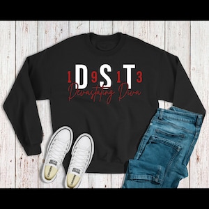 May include: Black sweatshirt with white text that reads "1D9ST13 Devastating Diva" in red lettering. The sweatshirt is laid out on a wood surface with a pair of white Converse sneakers and blue jeans.