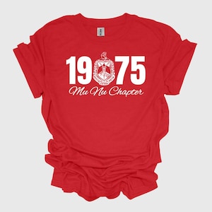 May include: A red t-shirt with white text and a crest. The text reads "1975" above "Mu Nu Chapter". The crest is a circular design with additional text. The shirt is made of a soft material.