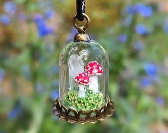 May include: A glass dome necklace with a miniature forest scene inside. The scene features three red and white mushrooms growing on a bed of green moss.
