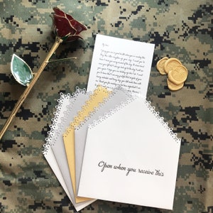 May include: Four white envelopes with scalloped edges, stacked on top of each other. The top envelope has the words "Open when you receive this" written in black ink. The envelopes are on a camouflage background with a gold-plated rose and three gold wax seals.