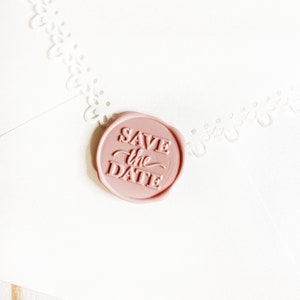 Könnte beinhalten: Ein rosa Wachssiegel mit dem geprägten Text "Save the Date".