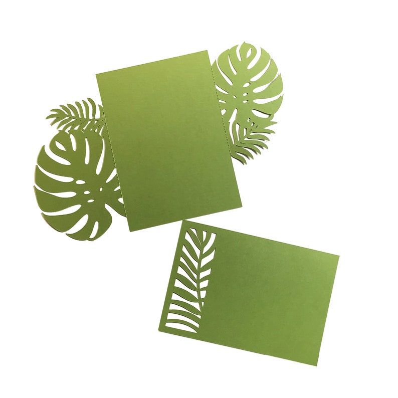 Puede incluir: Recortes de papel verde de hojas tropicales y dos tarjetas rectangulares. Las tarjetas son de color verde liso, y una tiene un dise&ntilde;o de hojas recortadas en el lateral. Las hojas tienen forma de hojas de monstera y helecho.