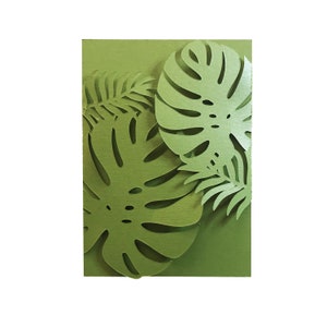 Puede incluir: Una tarjeta verde con recortes de papel en capas de hojas tropicales, incluyendo grandes hojas de monstera y follaje m&aacute;s peque&ntilde;o en forma de helecho. La tarjeta tiene una superficie texturizada.