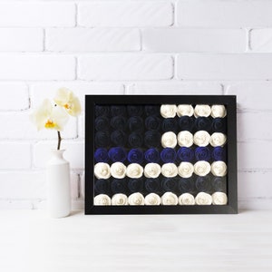 May include: A black framed wall art piece featuring a blue and white paper flower design. The flowers are arranged in a pattern resembling a flag.