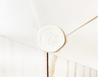 May include: A white envelope with a white wax seal featuring a bee design. The envelope is on a light wood surface.