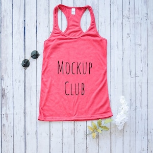 Puede incluir: Una camiseta sin mangas de color coral con las palabras "MOCKUP CLUB" en negro. Se muestra sobre una superficie de madera blanca, acompañada de gafas de sol negras y flores blancas.