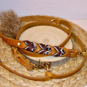 May include: A tan leather dog leash featuring a braided section with alternating dark brown leather and leopard print fabric accents. The leash includes gold-toned metal swivel snap hooks at both ends and a gold-toned rivet near the handle.