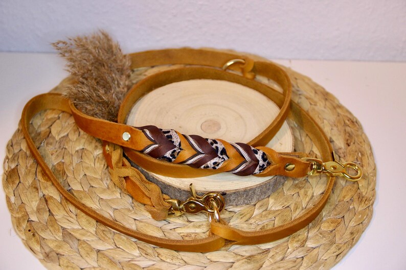 May include: A tan leather dog leash featuring a braided section with alternating dark brown leather and leopard print fabric accents. The leash includes gold-toned metal swivel snap hooks at both ends and a gold-toned rivet near the handle.